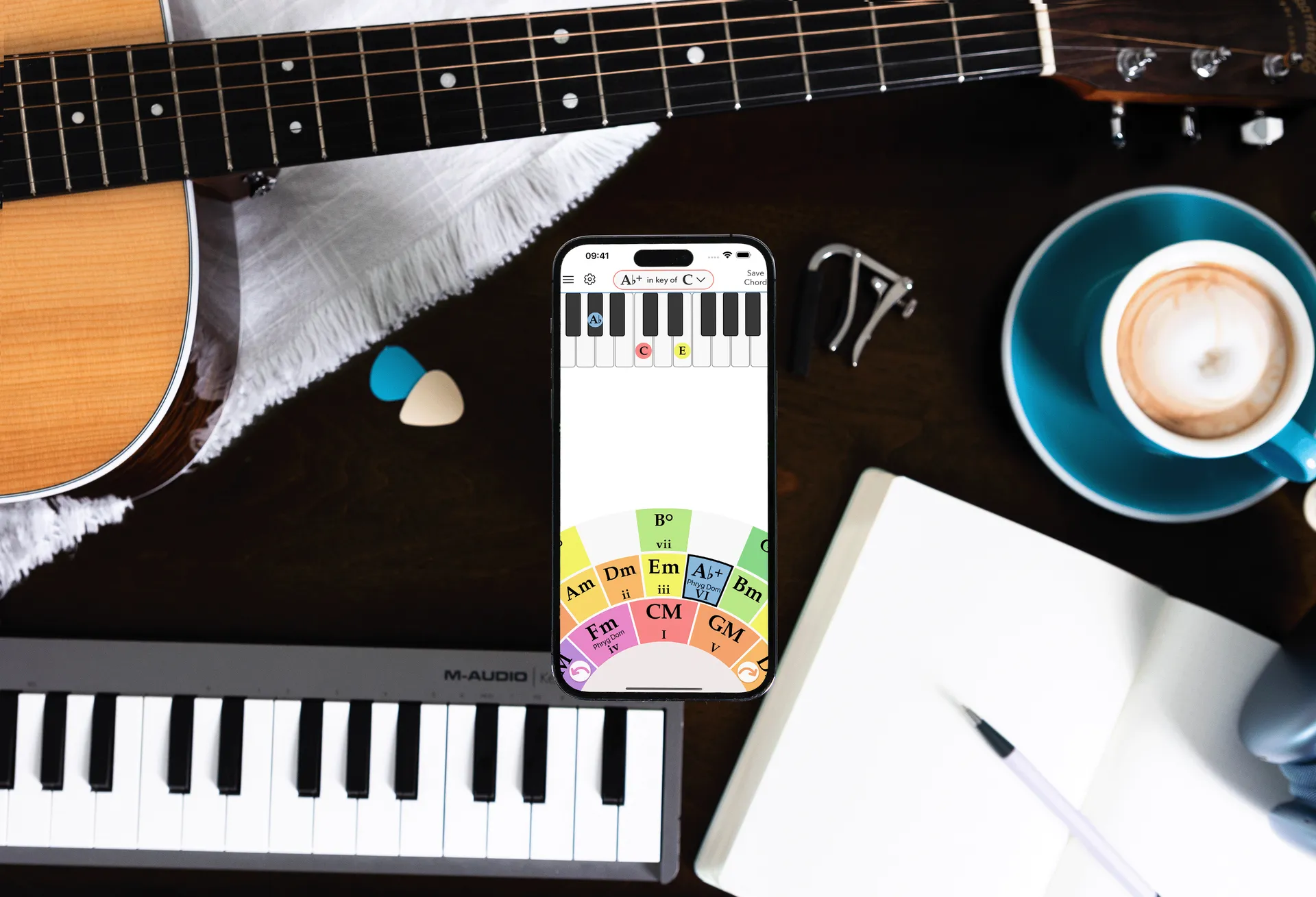 Chord Colors' interactive chord wheel on an iPhone, on a desk with an acoustic guitar, a MIDI keyboard, and a cup of coffee